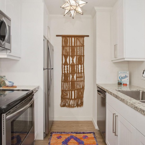 Apartments For Rent In Coronado, CA - Bayside At Coronado - Kitchen With Wood-Style Plank Flooring, Rug, Granite Countertops, White Cabinets, And Stainless-Steel Appliances.